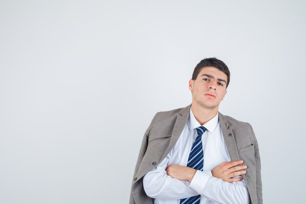 Ragazzo teenager in camicia, giacca, cravatta a righe in piedi con le braccia incrociate e guardando fiducioso, vista frontale.