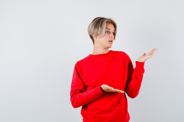 Ragazzo teenager in maglione rosso che finge di mostrare qualcosa e sembra sconcertato, vista frontale.