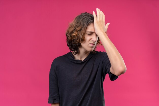 Ragazzo triste con i capelli lunghi in maglietta nera mise la mano sulla fronte sul muro rosa