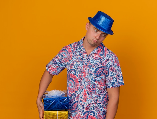 Ragazzo triste del partito giovane che indossa il cappello blu che tiene il contenitore di regalo isolato sull'arancio