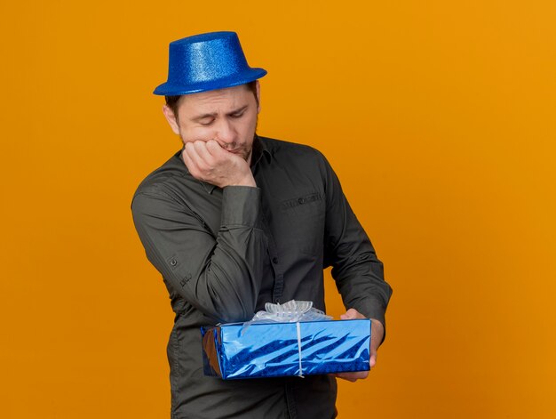 Ragazzo triste del partito giovane che porta la tenuta del cappello blu e che mette il gomito sul contenitore di regalo isolato sull'arancio