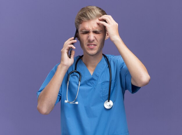 Rammaricato con gli occhi chiusi giovane medico maschio che indossa l'uniforme del medico con lo stetoscopio parla al telefono mettendo la mano sulla fronte isolata sulla parete blu