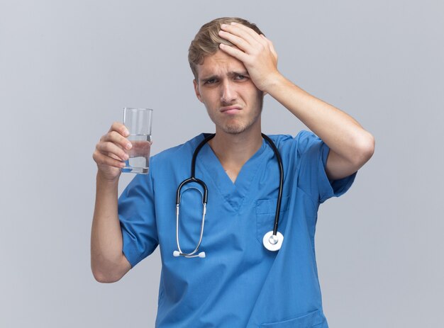 Rammaricato giovane medico maschio che indossa l'uniforme del medico con lo stetoscopio che tiene il bicchiere d'acqua mettendo la mano sulla testa isolata sul muro bianco