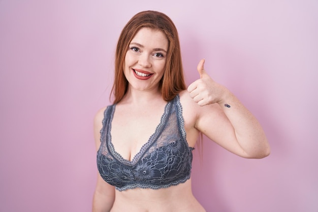 Redhead donna che indossa lingerie su sfondo rosa facendo felice pollice in alto gesto con la mano. espressione di approvazione guardando la telecamera che mostra il successo.