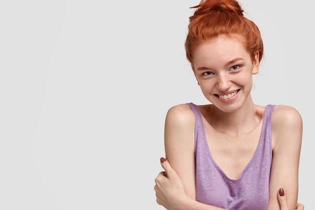 Redhead donna timida con la pelle lentigginosa, ha un sorriso gentile, tiene le mani incrociate sul petto, guarda con espressione gentile, ha una bella conversazione amichevole, sta contro il muro bianco, spazio libero