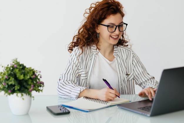 Redhead Ewoman contabile analizza i documenti utilizza la calcolatrice rende report utilizza il computer portatile si siede su un desktop bianco indossa occhiali maglione beige pone contro interni bianchi di casa