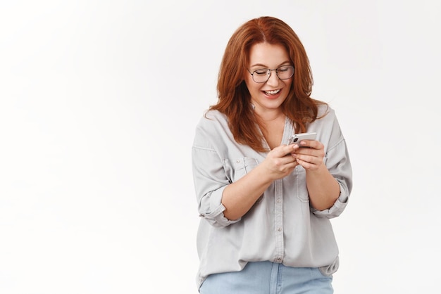 Redhead felice di mezza età moderno elegante donna SMS marito inviare emoji divertenti ridere leggere articolo divertente social media chat amici guardare smartphone display tenere telefono indossare occhiali muro bianco