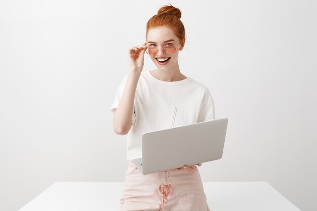 Redhead femminile alla moda in occhiali da sole che lavorano con il computer portatile