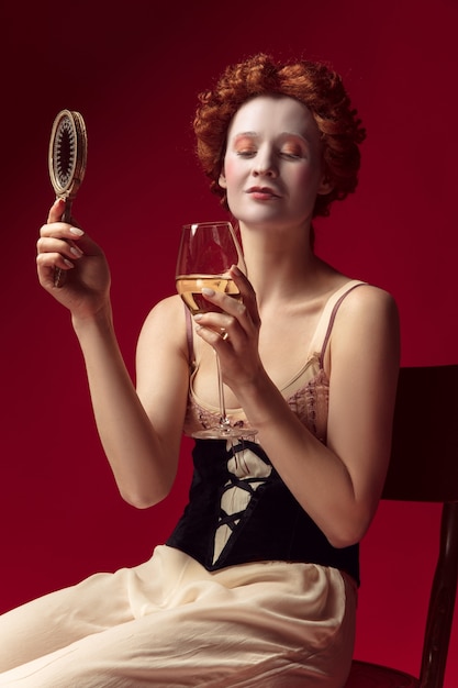 Redhead medievale giovane donna come una duchessa in corsetto nero e abiti da notte seduta su uno spazio rosso con uno specchio e un bicchiere di vino