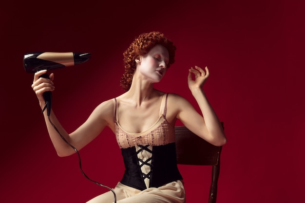 Redhead medievale giovane donna come una duchessa in corsetto nero e abiti da notte seduta sulla sedia sul muro rosso. Facendosi i capelli con l'asciugatrice. Concetto di confronto tra epoche, modernità e rinascita.