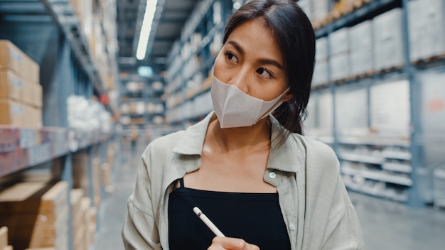 Responsabile della giovane donna di affari asiatica che indossa il magazzino della maschera per il viso utilizzando l'inventario di controllo della tavoletta digitale