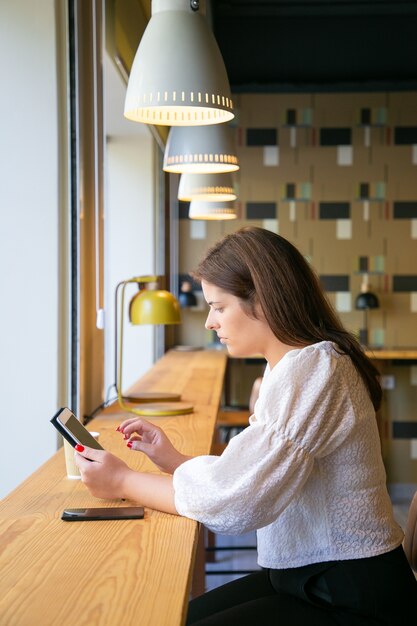 Responsabile femminile concentrato utilizzando tablet mentre è seduto alla scrivania in uno spazio di co-working o caffetteria