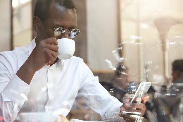 Responsabile nero bello nell'usura convenzionale che controlla email o che legge le notizie di mondo sul telefono cellulare digitale, bevendo cappuccino di mattina, sedendosi al tavolo alla caffetteria. Tecnologia, connessione e comunicazione