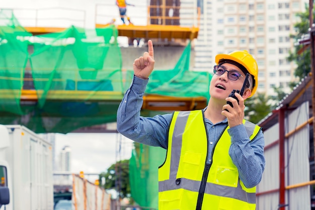 Responsabile tecnico asiatico intelligente con uniforme di sicurezza che controlla la costruzione del sito con sfondo di struttura in acciaio e cemento