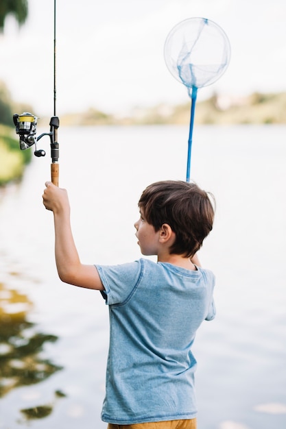 Retrovisione del ragazzo che solleva le mani che tengono canna da pesca e rete davanti al lago