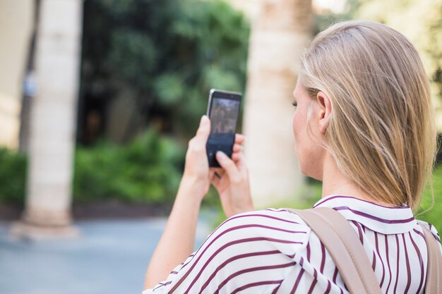 Retrovisione della giovane donna bionda che prende fotografia sul telefono cellulare