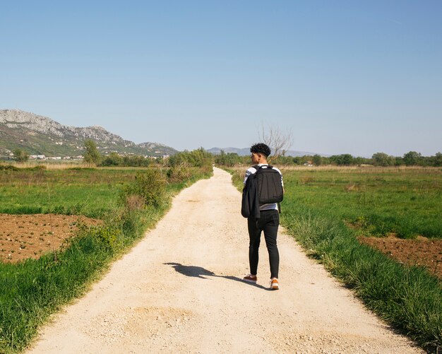 Retrovisione di giovane viaggiatore maschio che cammina nello zaino di trasporto del lato del paese