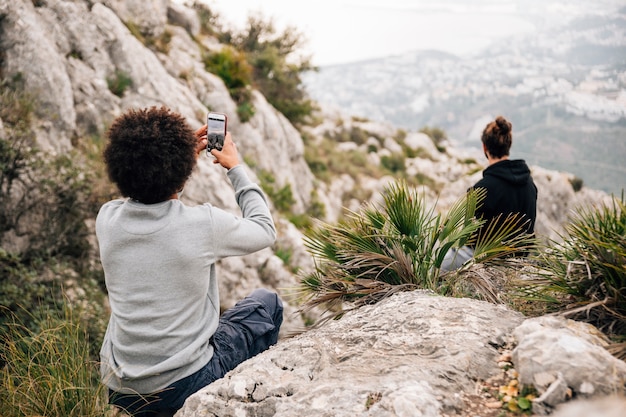 Retrovisione di un uomo che prende foto del suo amico che si siede sulla roccia con il telefono cellulare