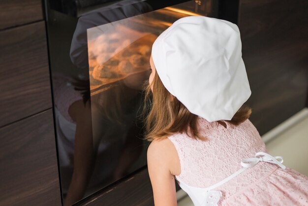 Retrovisione di una bambina in cappello del cuoco unico che esamina il vassoio del biscotto in forno