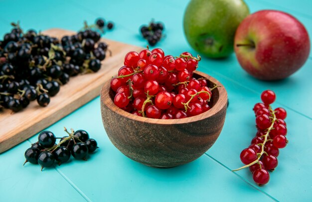 Ribes vista frontale in una ciotola con ribes nero su una tavola e mele su uno sfondo blu chiaro