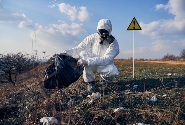 Ricercatore in tuta protettiva che raccoglie immondizia di plastica nel sacco nero dei rifiuti all'aperto in una giornata di sole