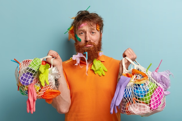 Riciclaggio e concetto di problema di inquinamento da plastica. Uomo rosso scontento con setole spesse, spazzatura conficcata nei capelli e nella barba, porta due sacchi pieni di immondizia, isolato sopra il muro blu