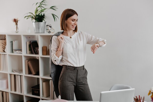 Ridendo donna d'affari in camicetta bianca e pantaloni grigi sta guardando l'orologio da polso, in piedi contro i mobili per ufficio.