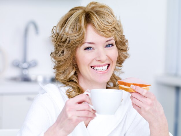 Ridendo giovane bella donna con una tazza di caffè facendo colazione - al chiuso