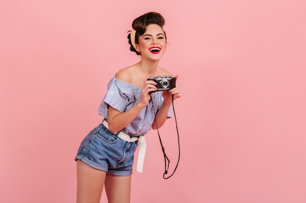 Ridendo giovane donna in shorts in denim a scattare foto. Studio shot di pinup girl con fotocamera isolata su sfondo rosa.