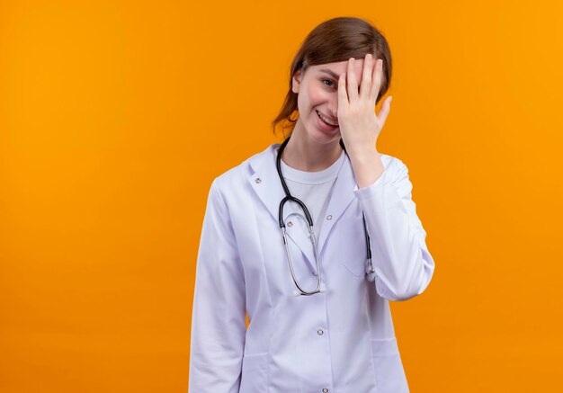 Ridendo giovane medico femminile che indossa abito medico e stetoscopio mettendo la mano sul viso sullo spazio arancione isolato con lo spazio della copia