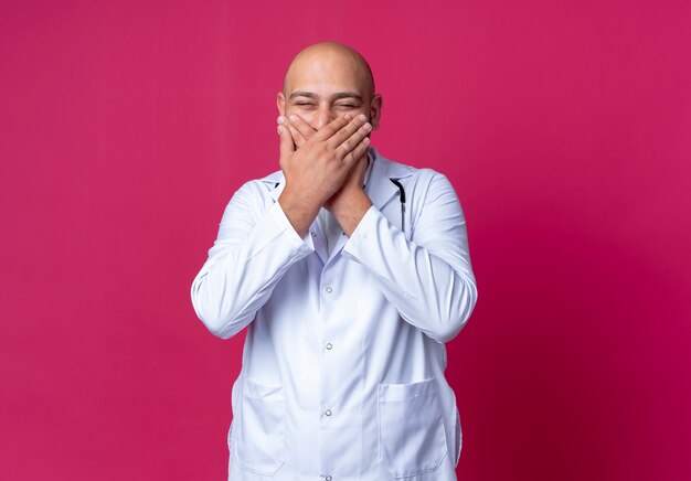 Ridendo giovane medico maschio calvo che indossa abito medico e stetoscopio ha coperto la bocca con la mano isolata sulla parete rosa