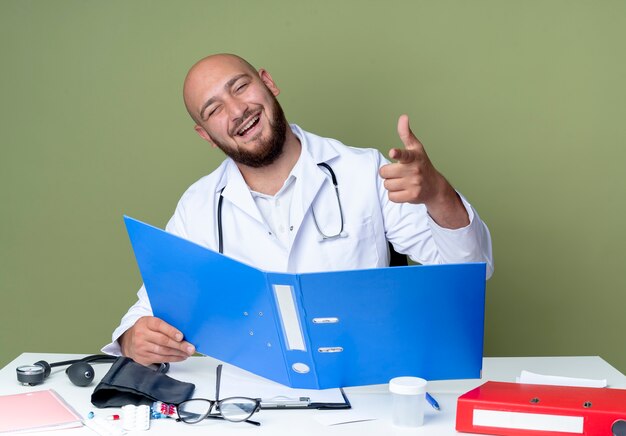 Ridendo giovane medico maschio calvo indossando accappatoio medico e stetoscopio seduto alla scrivania