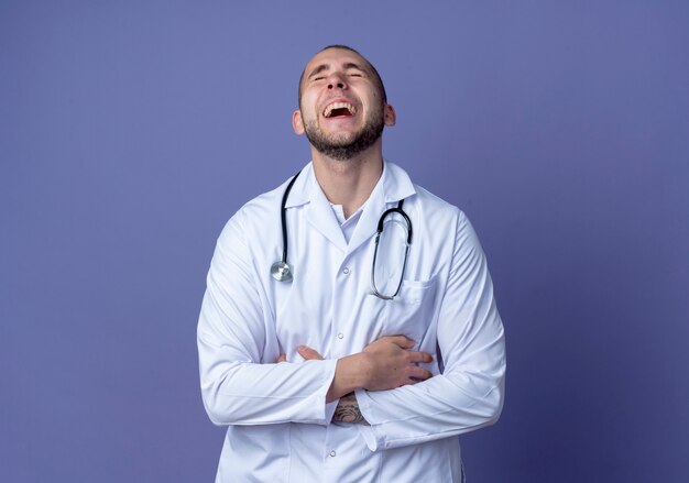 Ridendo giovane medico maschio che indossa veste medica e stetoscopio mantenendo le mani incrociate sulla pancia con gli occhi chiusi isolato su sfondo viola con spazio di copia