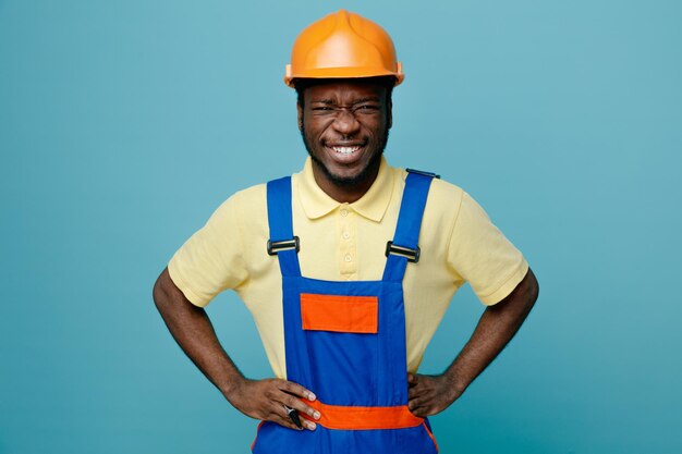 Ridendo mettendo le mani sui fianchi giovane costruttore afroamericano in uniforme isolato su sfondo blu