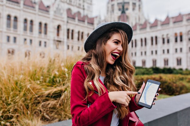 Ridendo splendida donna in cappello che mostra lo schermo del telefono mentre esplora la parte vecchia della città