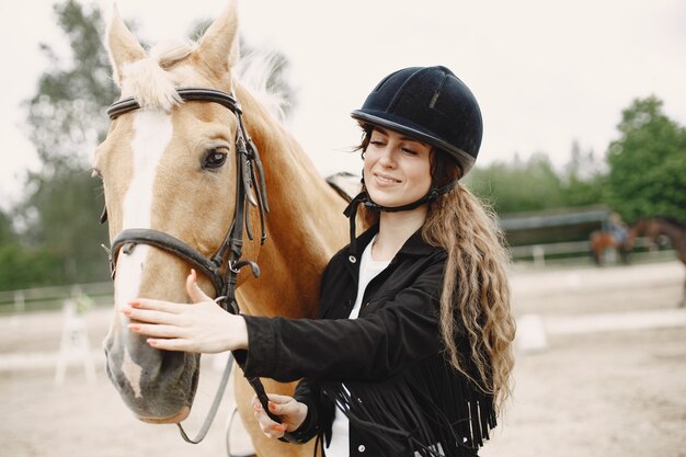 Rider donna che parla con il suo cavallo in un ranch. La donna ha i capelli lunghi e vestiti neri. Equestre femminile che tocca il suo cavallo marrone.