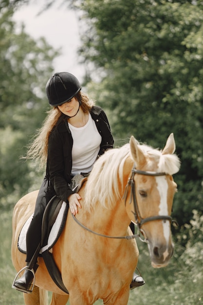 Rider donna in sella al suo cavallo in un ranch. La donna ha i capelli lunghi e vestiti neri. Equestre femminile che tocca il suo cavallo marrone.