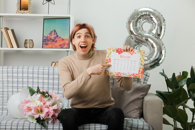 Ridere bel ragazzo durante la giornata delle donne felici che tiene e indica il calendario seduto sul divano nel soggiorno