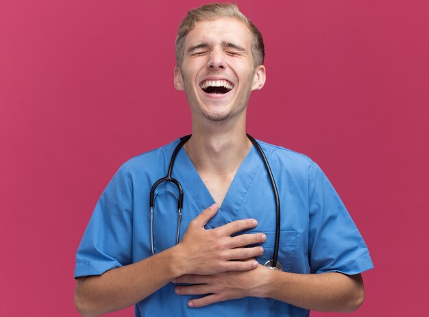 Ridere con gli occhi chiusi giovane medico maschio che indossa l'uniforme del medico con lo stetoscopio isolato sulla parete rosa