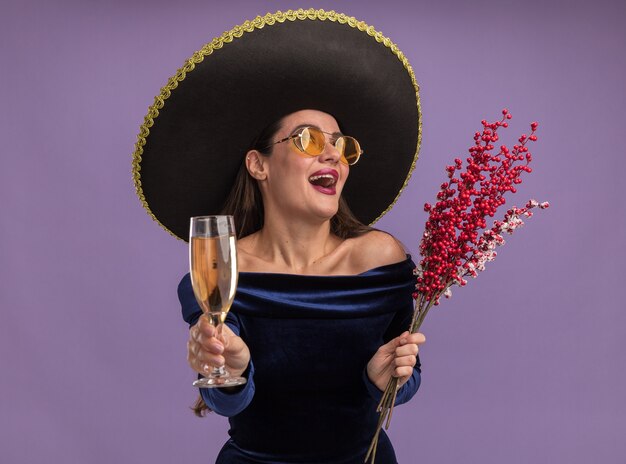 Ridere giovane bella ragazza che indossa un abito blu e bicchieri con sombrero che tiene il ramo di sorbo con un bicchiere di champagne isolato su sfondo viola