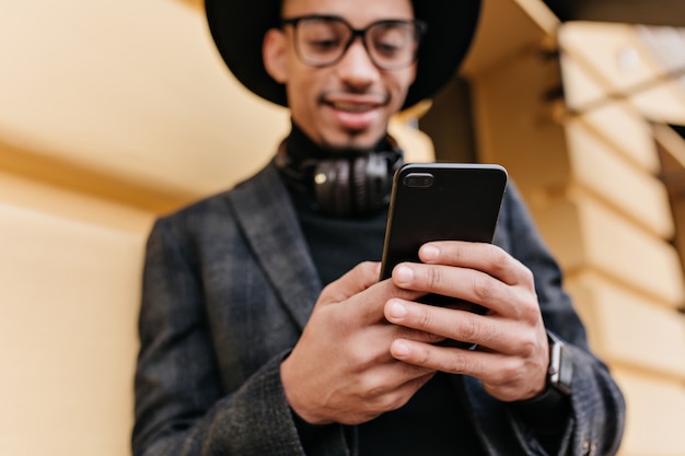 Ridere il messaggio di sms di uomo mulatto sulla strada urbana. Ritratto di sfocatura all'aperto del ragazzo sorridente africano con il telefono a fuoco.