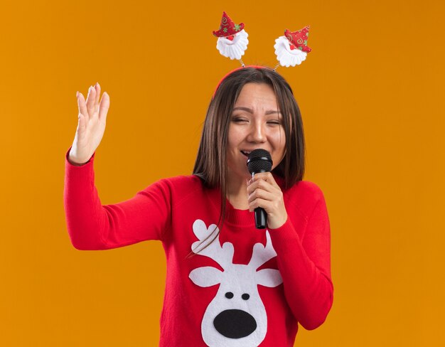 Ridere la giovane ragazza asiatica che indossa il cerchio dei capelli di Natale con il maglione parla al microfono isolato sulla parete arancione