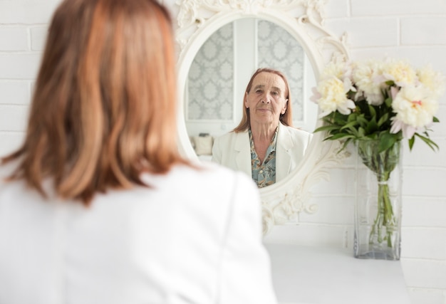 Riflessione della donna senior sullo specchio vicino al bello vaso di fiore a casa
