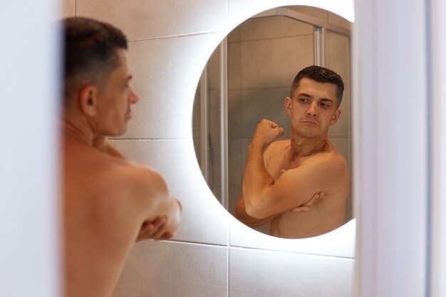 Riflessione nello specchio uomo fiero sicuro di sé con i capelli scuri e il corpo nudo, guardando i suoi bicipiti con espressione facciale soddisfatta, in posa in bagno.