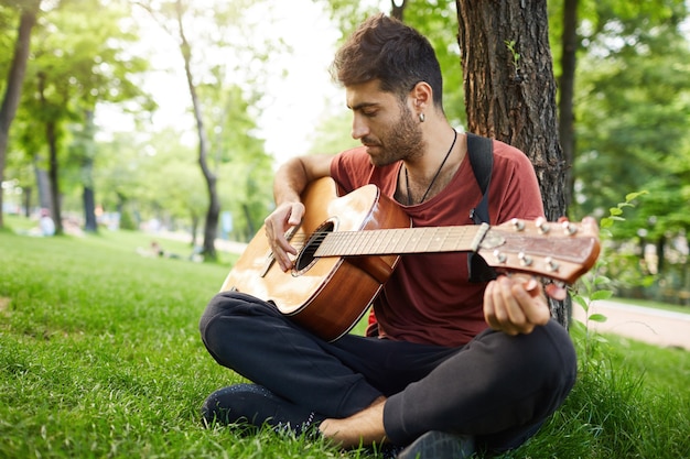 Riflessivo bel giovane uomo suonare la chitarra al parco, appoggiato su un albero e sedersi sull'erba