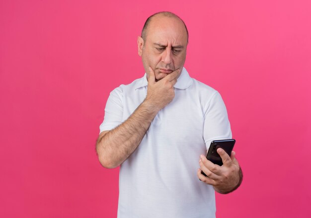 Riflessivo casual imprenditore maturo tenendo e guardando il telefono cellulare e tenendo la mano sul mento isolato su sfondo rosa con copia spazio
