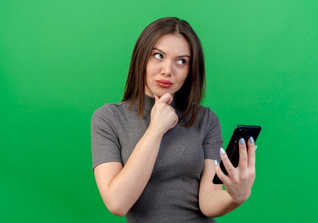 Riflessivo giovane donna graziosa che tiene il telefono cellulare toccando il mento e guardando il lato isolato su sfondo verde con spazio di copia