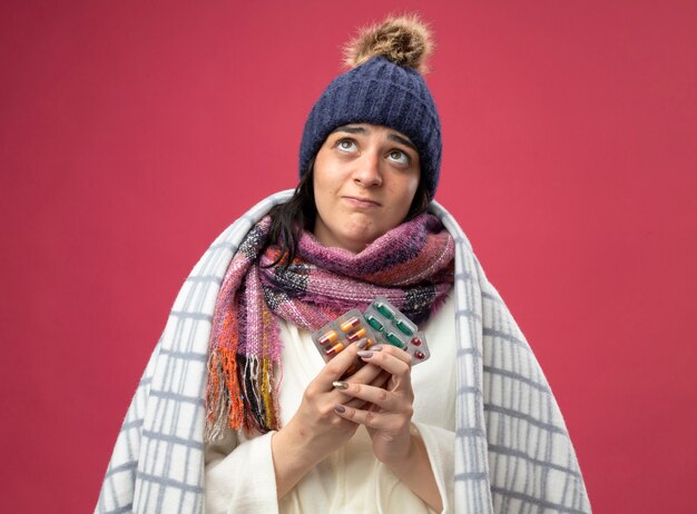 Riflessivo giovane indoeuropea ragazza malata che indossa abito invernale cappello e sciarpa avvolti in plaid che tiene confezioni di capsule cercando isolato sulla parete cremisi