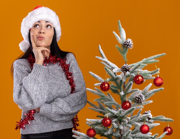 Riflessivo giovane ragazza caucasica indossando il cappello di Natale e la ghirlanda di orpelli intorno al collo in piedi vicino all'albero di Natale decorato tenendo la mano sul mento guardando in alto isolato su sfondo arancione