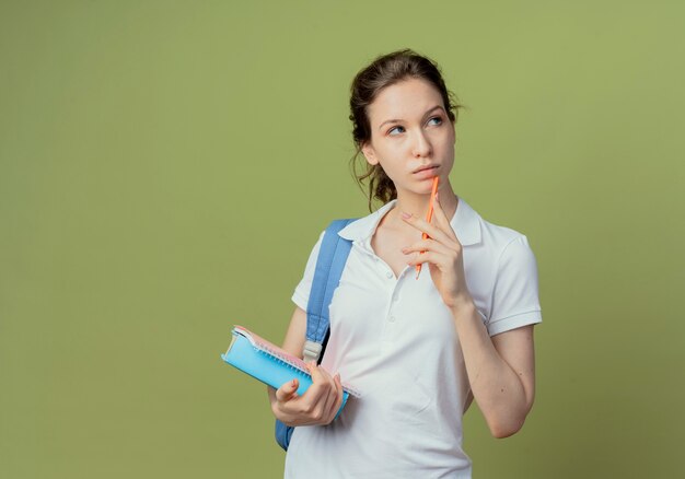 Riflessivo giovane studentessa graziosa che indossa la borsa posteriore guardando il lato tenendo appunti e libro e toccando il mento con la penna isolata su sfondo verde oliva con spazio di copia